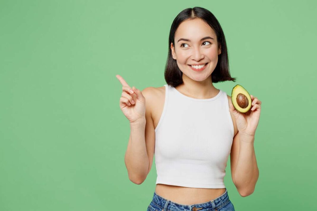 Neteisingas avokado valgymas – jauna moteris rankoje laiko avokadą ir šypsosi parodydama į šalį.