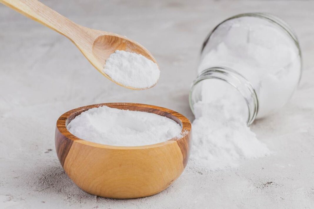 Soda kūno detoksikacijai – baltas miltelis mediniame indelyje su šaukštu ant šviesaus paviršiaus.