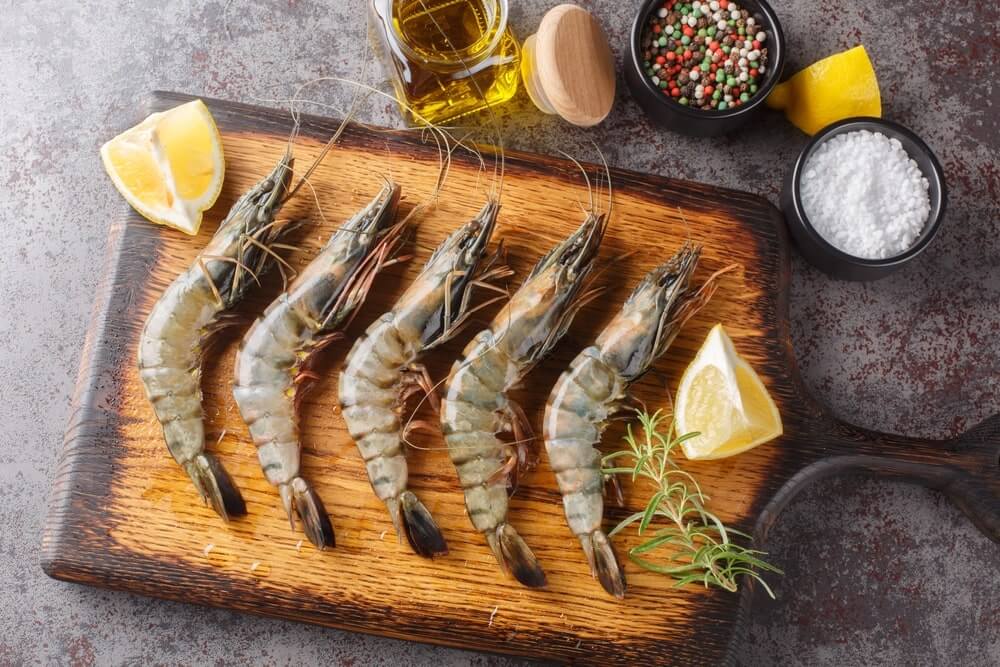 Ar sveika valgyti krevetes – žalios krevetės ant medinės lentos su citrina, aliejumi ir prieskoniais, paruoštos gaminimui.