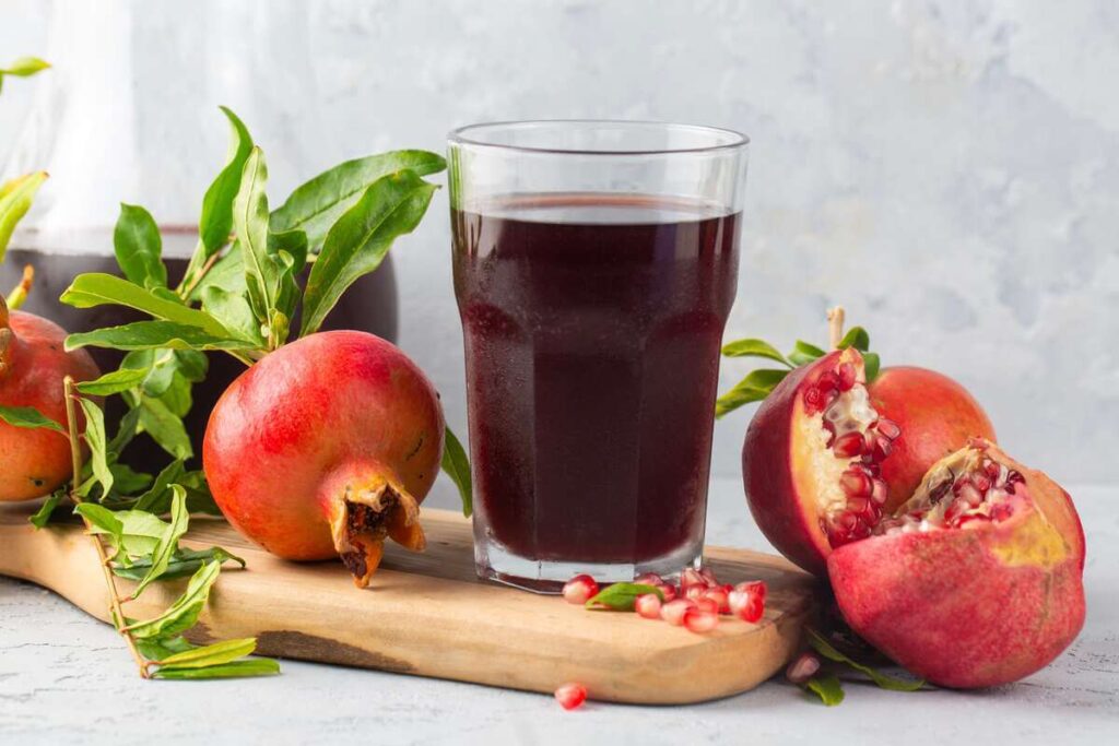 Granatų sultys aukštam spaudimui stiklinėje, apsuptoje prinokusių granatų ir sėklų.