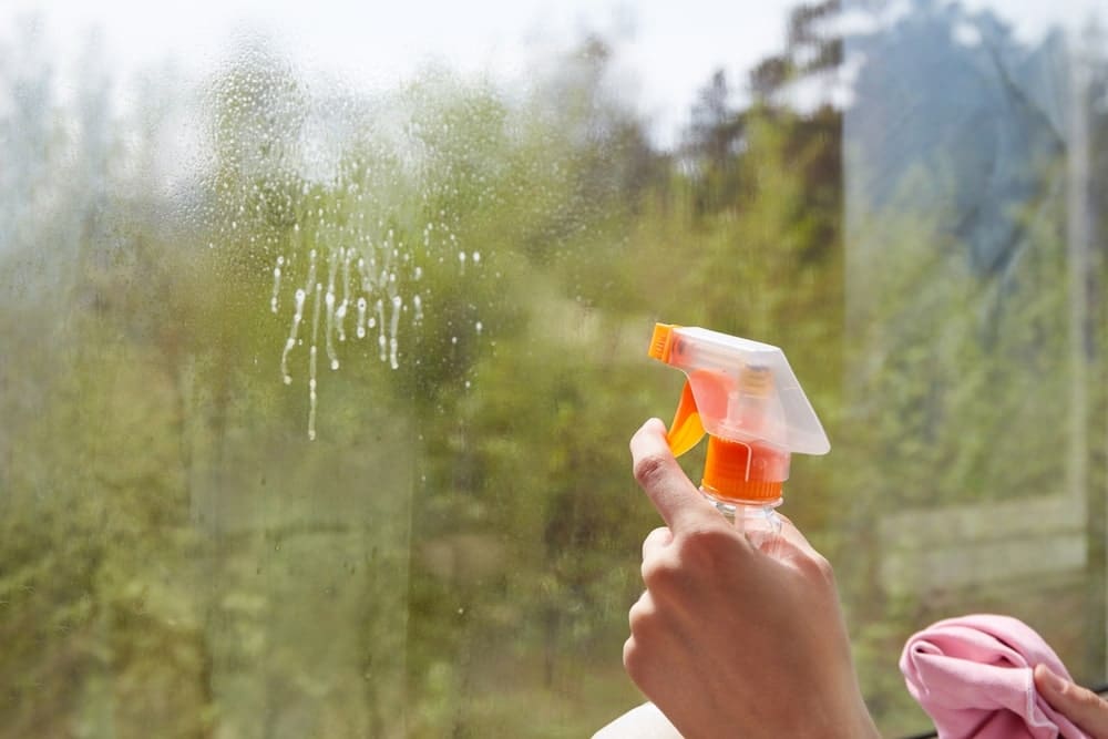 Indų ploviklis stiklų valymui naudojamas purškiant tirpalą ant lango – matomi vandens lašai ir valomas stiklo paviršius.
