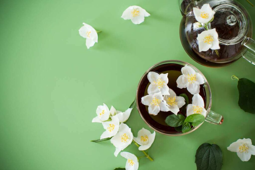 jazminų arbatos paruošimas su puodeliu ir baltais jazminų žiedais ant žalio fono, kuriant gaivų pavasario įspūdį.