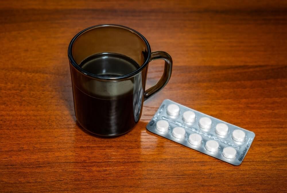 Kava ir vaistai – juodos kavos puodelis ir vaistų lizdinė plokštelė ant stalo, primenantys apie vartojimo laiką.