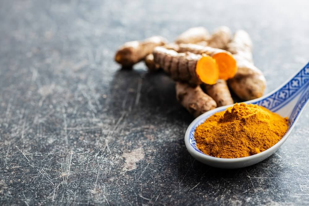 Kiek laiko ciberžolė išlieka organizme – ciberžolės milteliai šaukštelyje šalia šviežios šaknies, vaizduojant natūralią vartojimo formą.