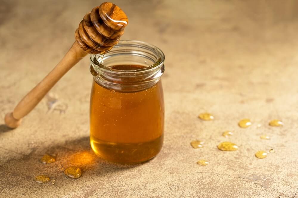 medaus nauda ir žala vaizduojama stiklainiui su medumi ir mediniu samteliu ant šviesaus paviršiaus