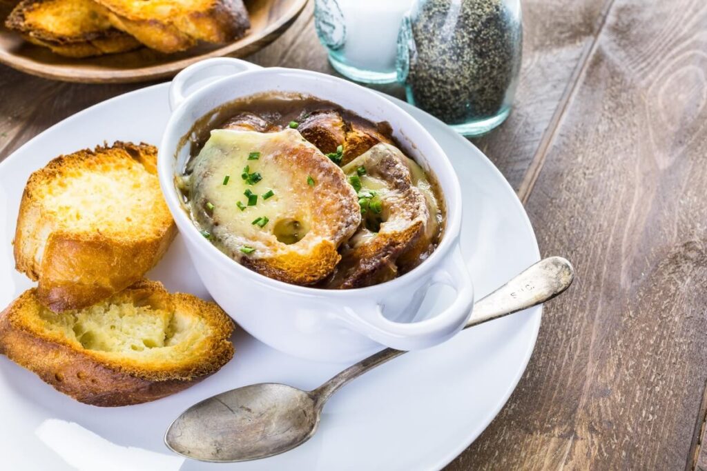 Prancūziška svogūnų sriuba dubenyje su apskrudusia bagete ir tirpstančiu sūriu, patiekta su papildomomis riekelėmis.