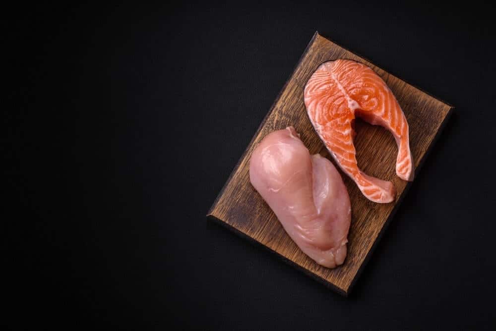 Vištiena ar lašiša – žalia vištienos krūtinėlė ir lašišos kepsnys ant medinės pjaustymo lentos.