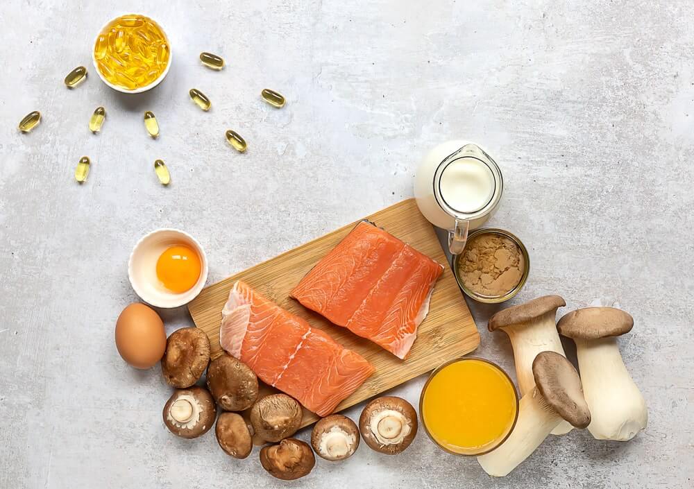 vitamino D šaltiniai maiste išdėstyti ant stalo – riebi žuvis, kiaušiniai, grybai ir pieno produktai