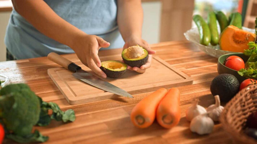 avokadas po 60, rankos pjausto prinokusį avokadą virtuvėje ant medinės lentos