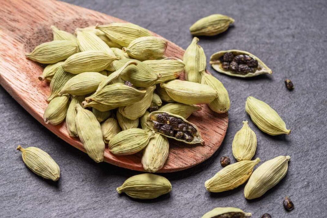 Kardamonas virtuvėje – žalios ankštys ir sėklos ant medinės lentelės, aiškiai matoma prieskonio tekstūra.