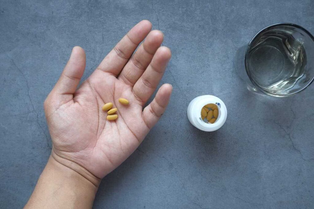 vitaminai ryte, tabletės laikomos delne šalia stiklinės vandens ant pilko paviršiaus