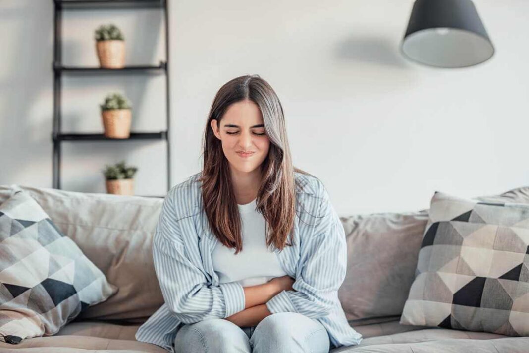 žarnyno veiklos sutrikimai atsispindi moters veide ir laikysenoje sėdint ant sofos namų aplinkoje