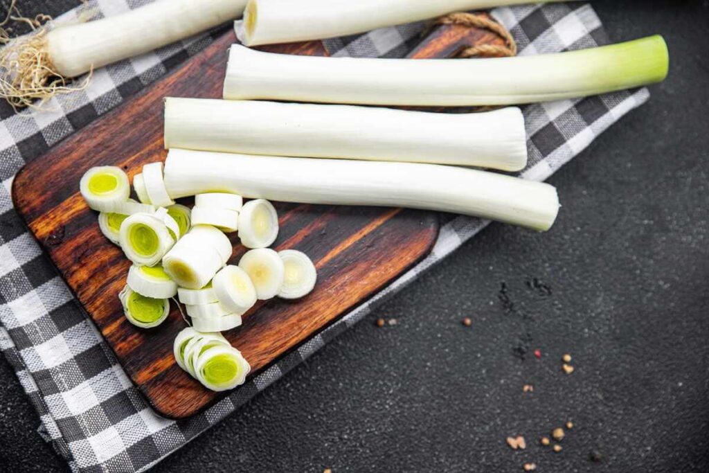 Daržiniai porai supjaustyti baltais griežinėliais ant medinės lentelės šalia sveikų stiebų.