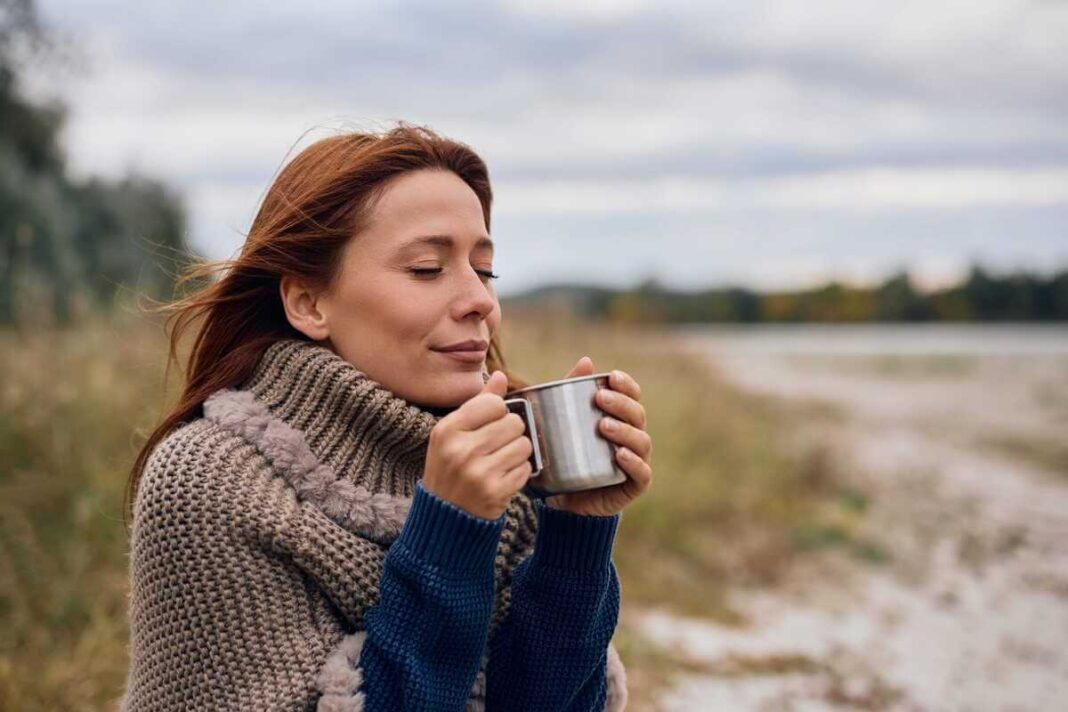 juodoji arbata metaliniame puodelyje, kurį laiko gamtoje atsipalaidavusi moteris užmerktomis akimis.