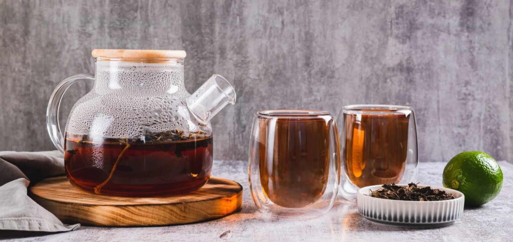 juodoji arbata skaidriame arbatinuke ir dviejuose puodeliuose ant stalo šalia žaliosios citrinos.