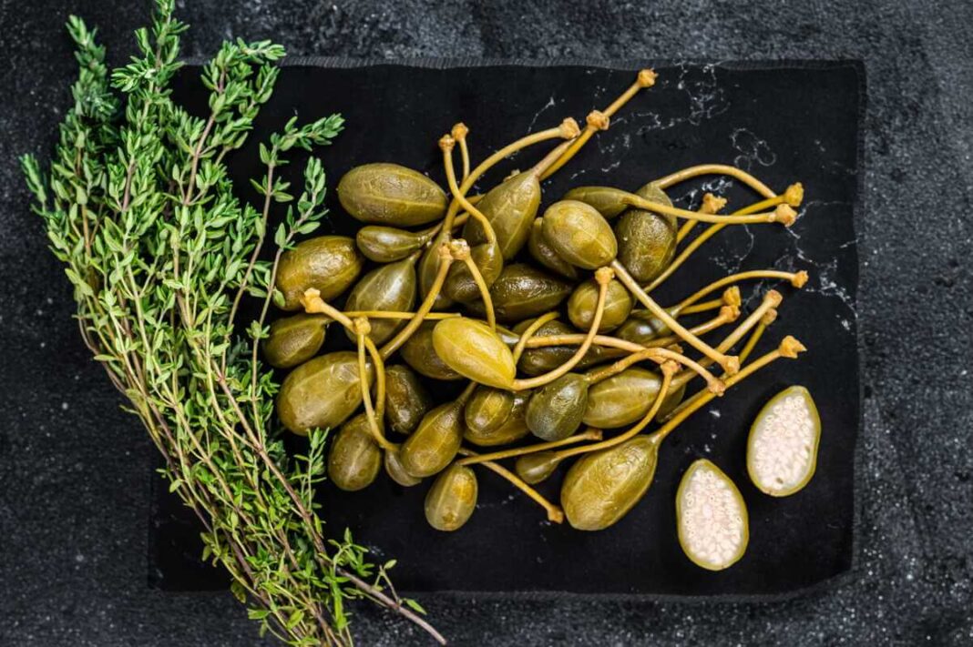 Kaparėlių nauda sveikatai iliustruojama vaizduojant marinuotus pumpurus su ilgais koteliais šalia šviežių čiobrelių šakelių.