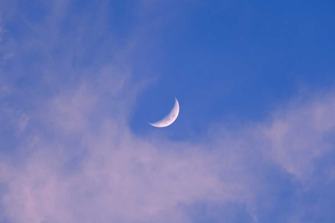 Mėnulio kalendorius su matomu jaunu pjautuvu tarp šviesių debesų giedrame dienos danguje.