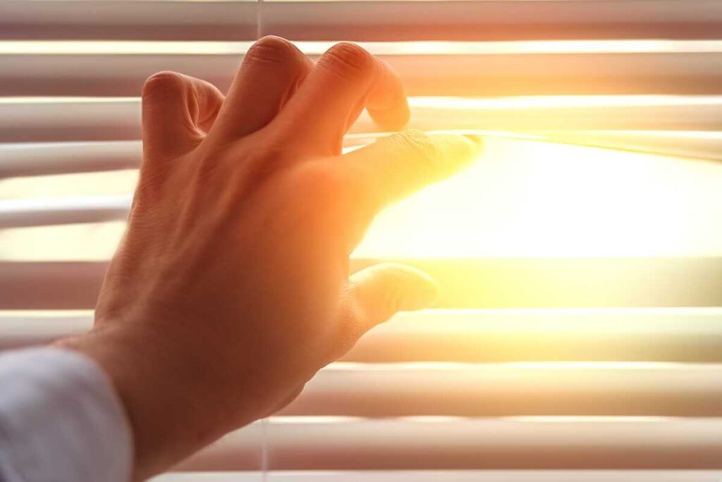 vitamino D trūkumas po žiemos saulės šviesa pro žaliuzes ir ranka, siekianti dienos šviesos