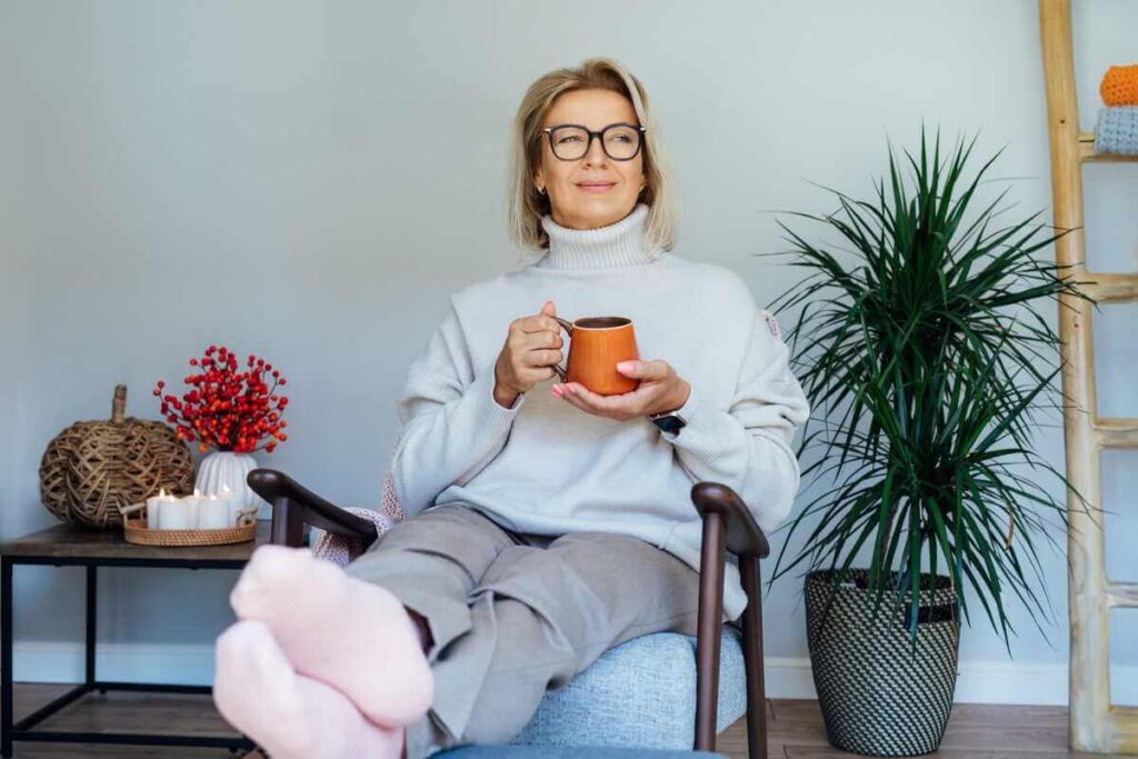Kavos nauda sveikatai ir atsipalaidavimui, kai moteris su akiniais sėdi fotelyje laikydama molinį puodelį šviesiame kambaryje.