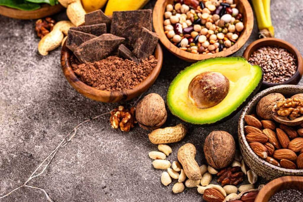 Magnio turintys produktai: ant pilko paviršiaus išdėlioti dubenėliai su riešutais, sėklomis, kakava ir perpjautas avokadas.