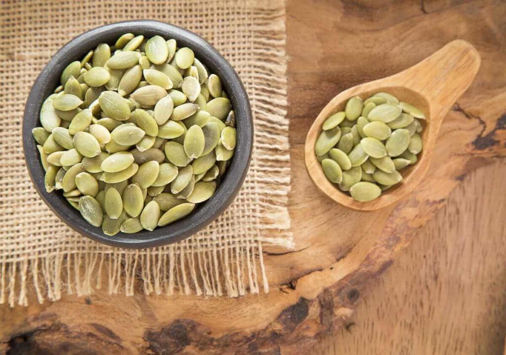 Moliūgų sėklos pavaizduotos pilname tamsiame dubenėlyje ir mediniame šaukšte, padėtuose ant natūralaus medžio paviršiaus su džiuto audiniu.