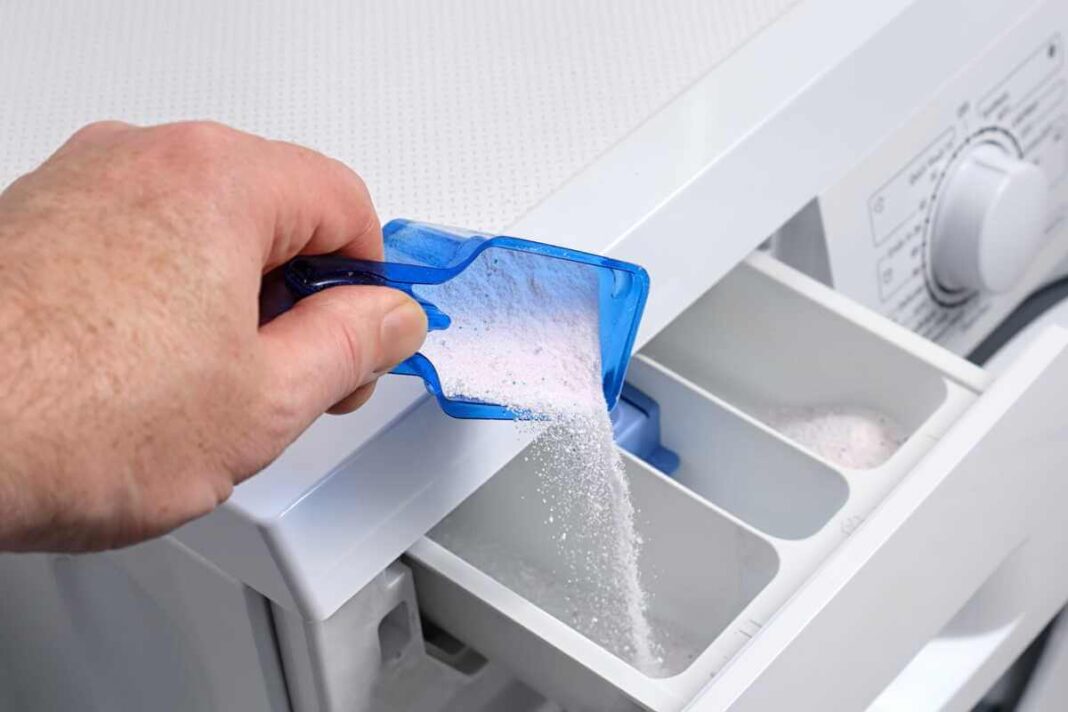 Skalbimo milteliai baltiems drabužiams: vaizduojamas atidarytas skalbimo mašinos stalčiukas, į kurį iš mėlyno plastikinio dozatoriaus pilami balti skalbimo milteliai.