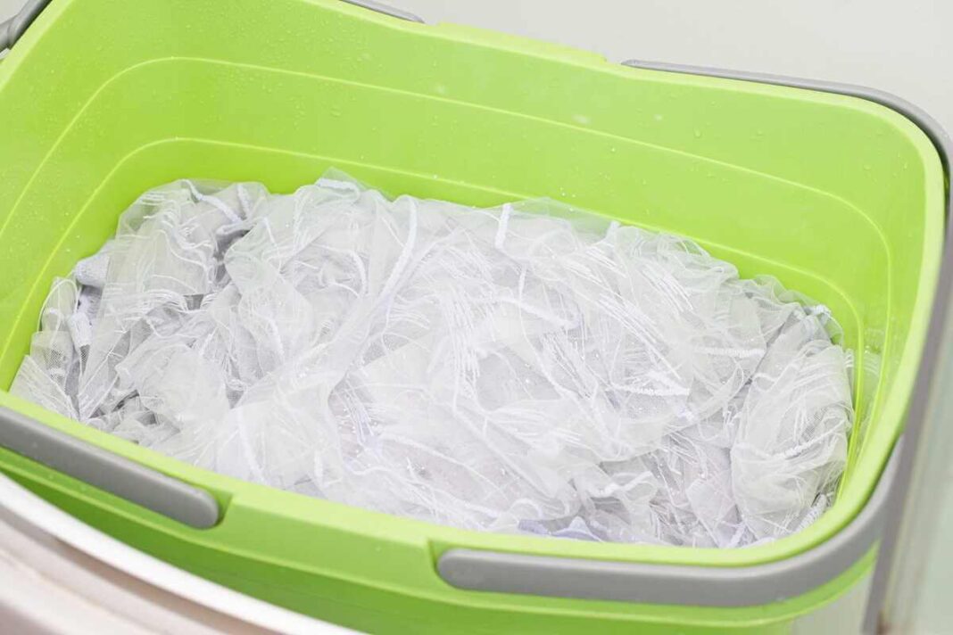 Užuolaidų balinimas namuose: skaidri tūlio užuolaida pamerkta į šiltą vandenį su natūraliomis priemonėmis žaliame plastikiniame dubenyje.