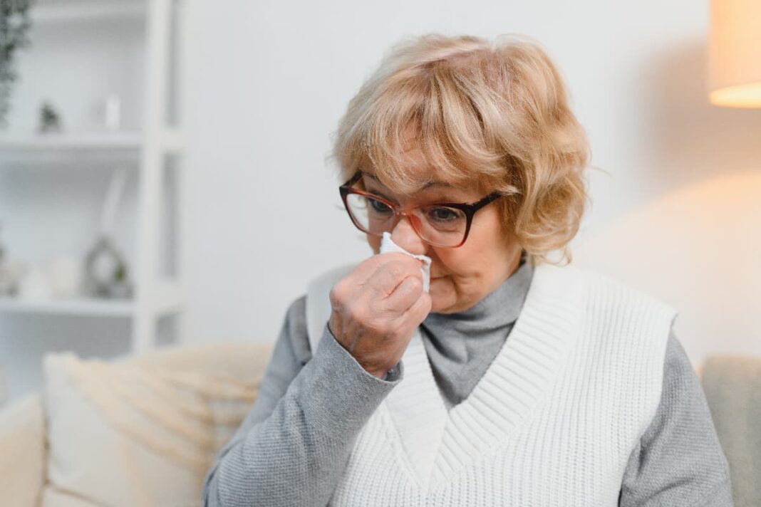 Vyresnė moteris akiniais prisidengia nosį ir burną balta servetėle