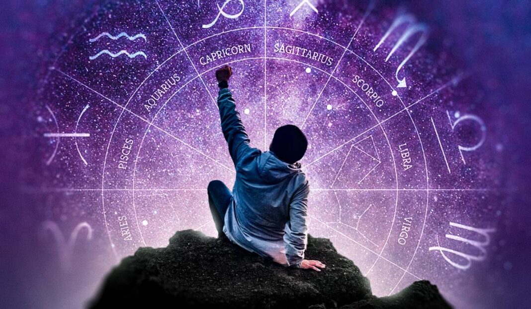 Žmogus ant uolos ranka siekia švytinčio Zodiako rato violetiniame kosmose